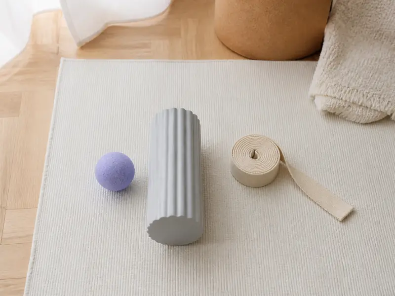 Balle de massage lilas, rouleau de mousse gris clair et sangle de coton écru alignés sur un tapis crème