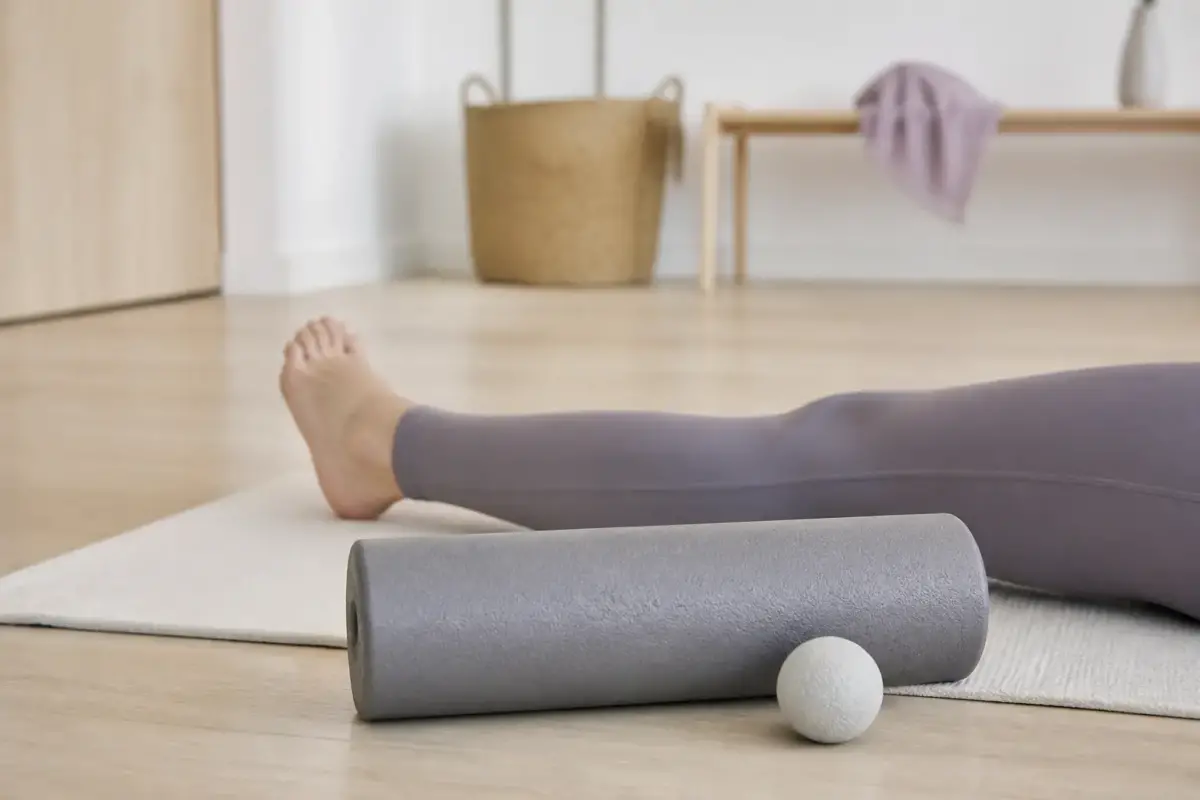 Rouleau en mousse gris et petite balle posés sur un tapis de sol, près d’une jambe allongée
