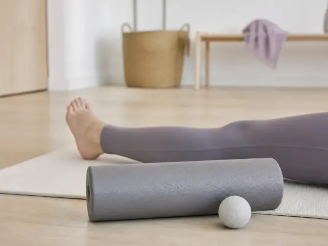 Rouleau en mousse gris et petite balle posés sur un tapis de sol, près d’une jambe allongée