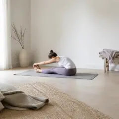 Personne assise au sol, jambes tendues devant elle, mains glissant vers les chevilles, vue de profil lointain