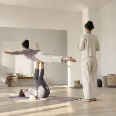 Deux personnes vues de loin en figure d’acroyoga, l’une allongée au sol soutenant l’autre sur ses pieds, une troisième à côté